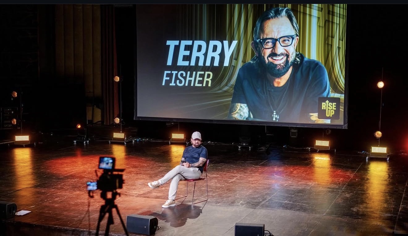 Terry Fisher seated on stage beneath a giant screen showing his name at RISE UP London